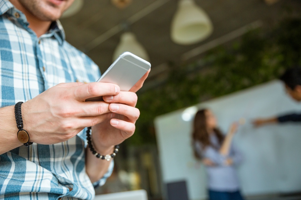 Closeup Of Hands Of Young Man In Checkered Shirt Using Mobile Phone While His Partners Arguing